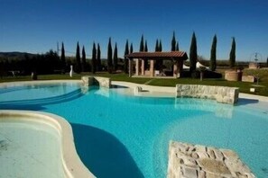 A swimming pool with a stone structure in the center, surrounded by a well-maintained lawn and tall trees. - La Pia Dama - Residence (Sinalunga)