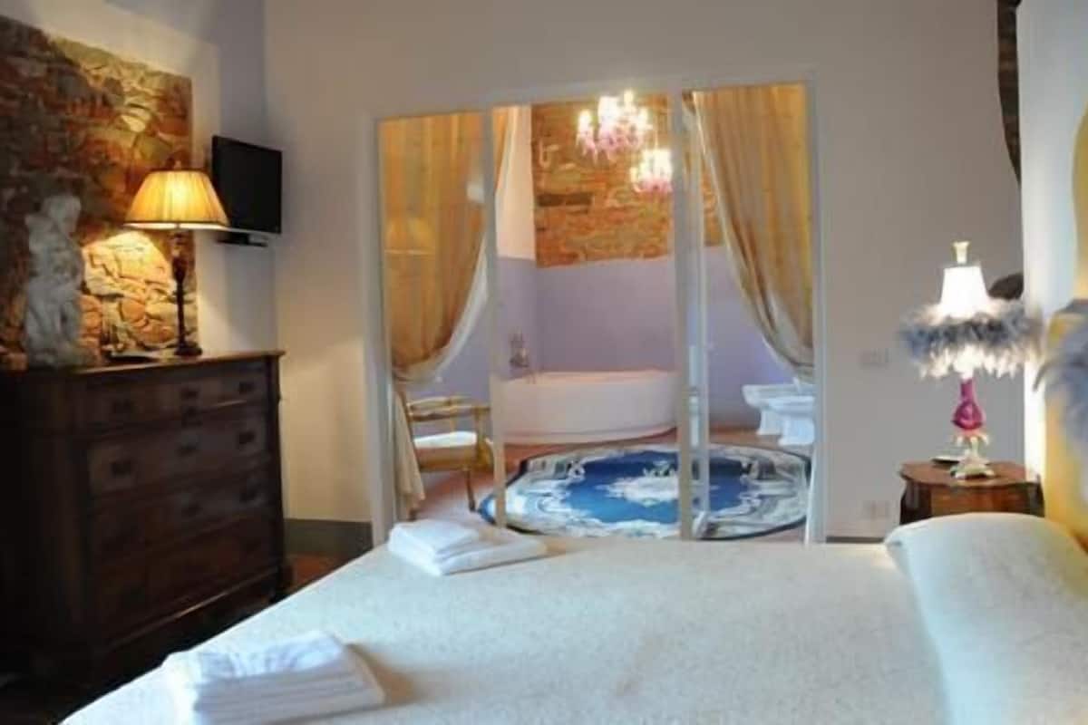 Une chambre à coucher comprenant un lit, une commode, un téléviseur, un lustre, et une salle de bain visible à travers une porte en verre.