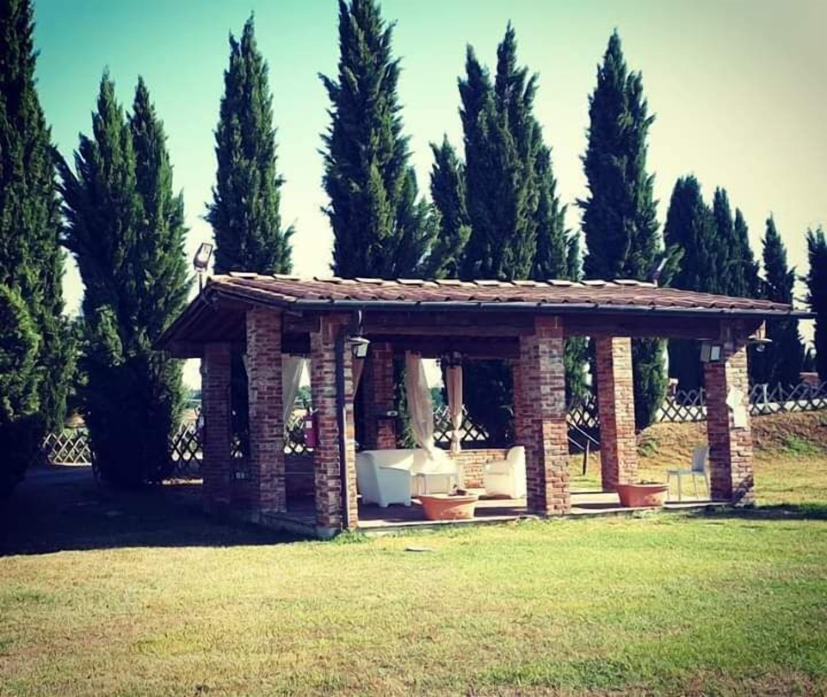 A brick pavilion with a tiled roof, surrounded by grass and tall trees.