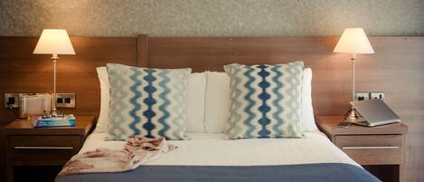 Una cama bien hecha con un edredón azul, dos almohadas estampadas y dos lámparas de noche. Hay dos mesitas de noche, una con una computadora portátil y la otra con una lámpara y un libro.