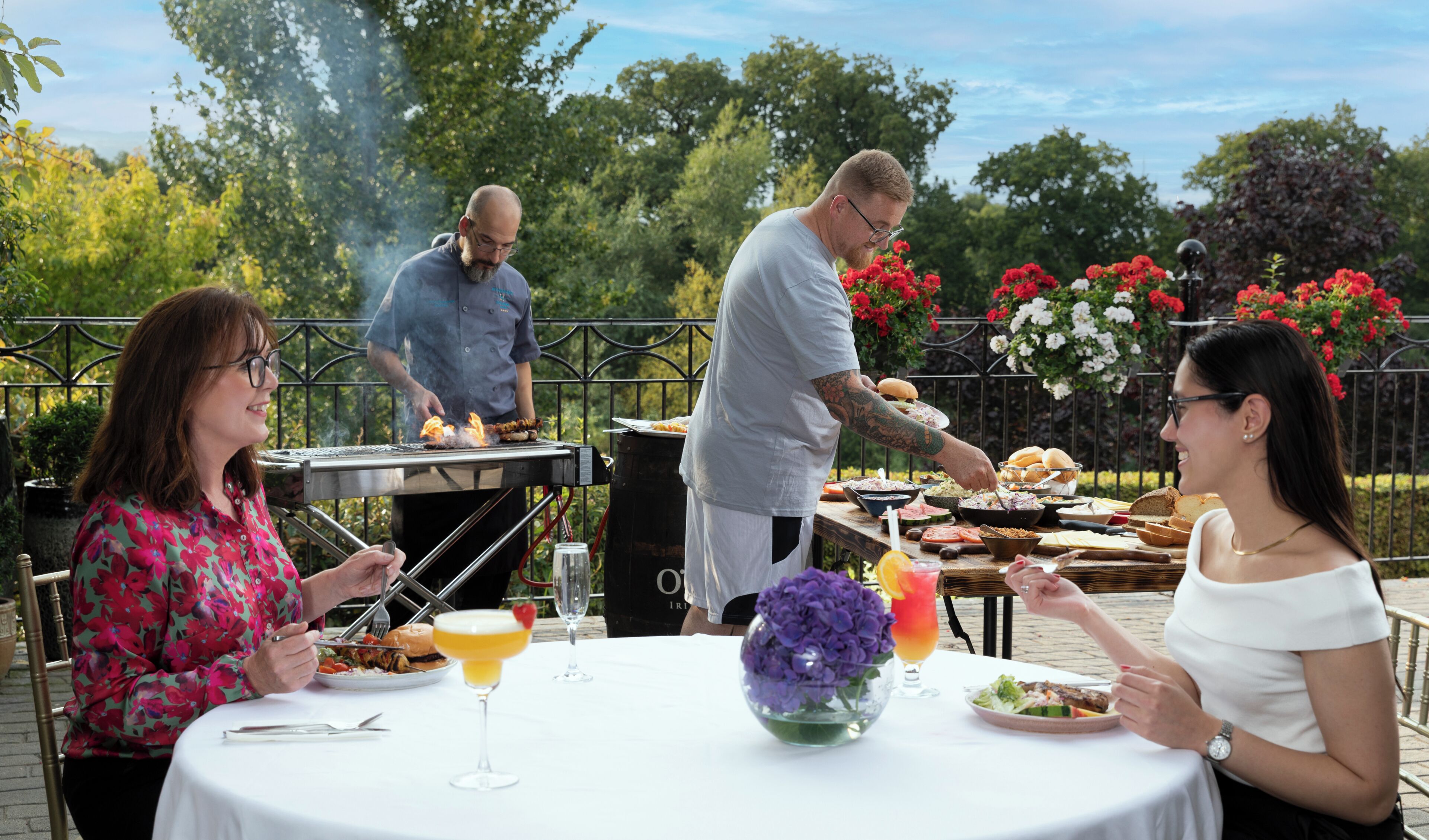An outdoor dining area with a grill, tables, and a view of trees and flowers.