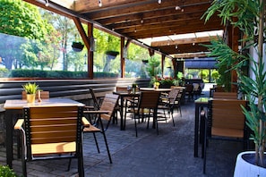 An outdoor cafe with wooden tables and chairs, potted plants, and a view of greenery through large windows. - Woodford Dolmen Hotel (Carlow)