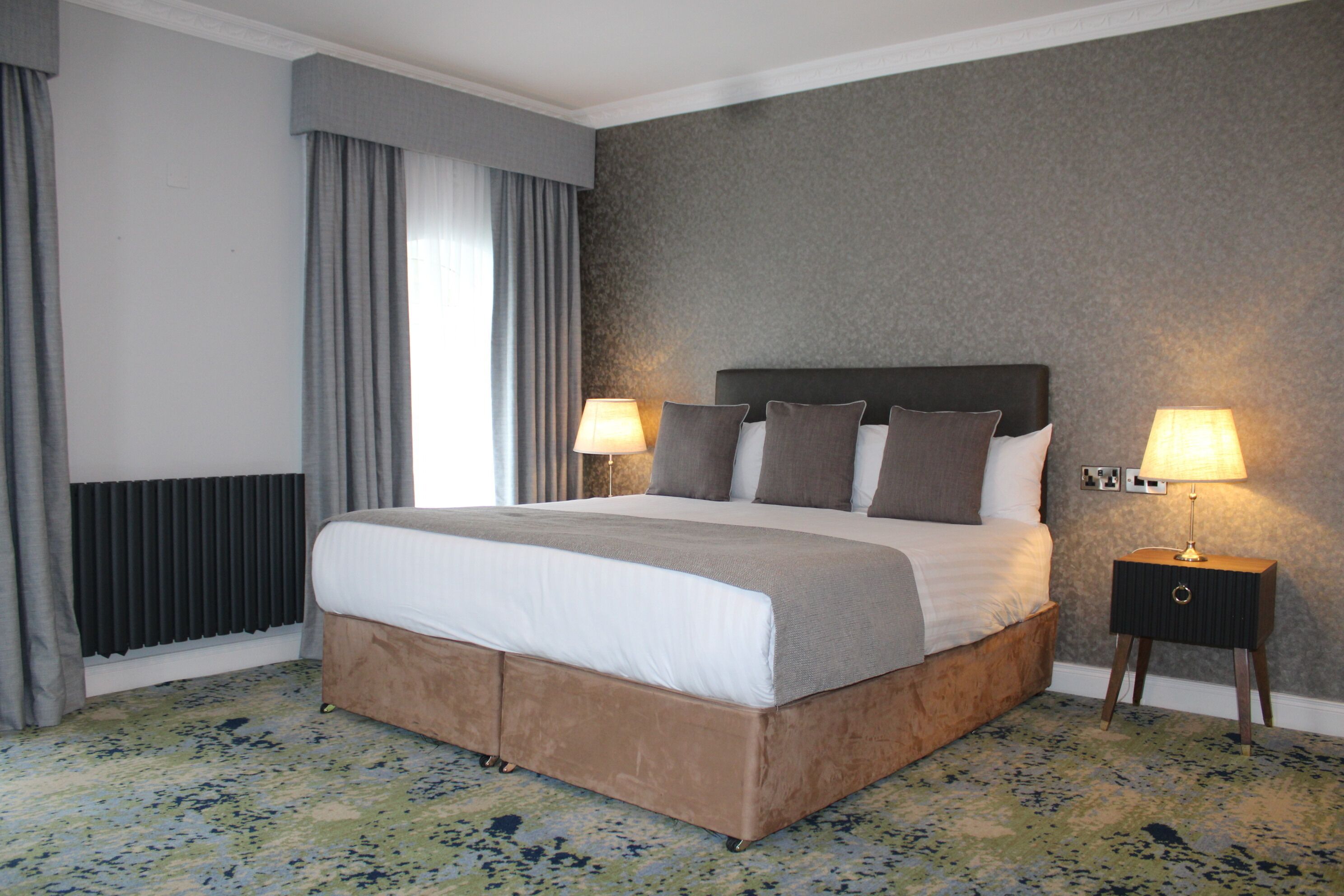 Habitación de hotel con una cama grande, dos lámparas de noche, un radiador y una alfombra estampada.