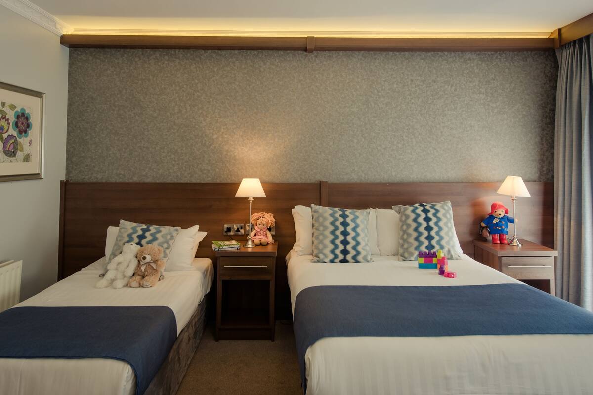 Ein Hotelzimmer mit zwei Betten, einem Kopfteil, Nachttischen, Lampen, einem Teddybären und einem gerahmten Bild an der Wand.