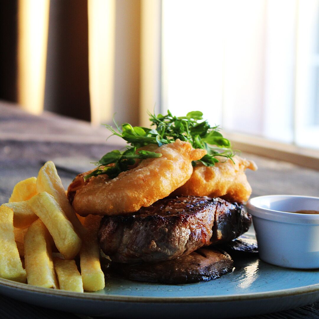 Un plato con un bistec, dos porciones de pescado frito, papas fritas y un recipiente pequeño de salsa.