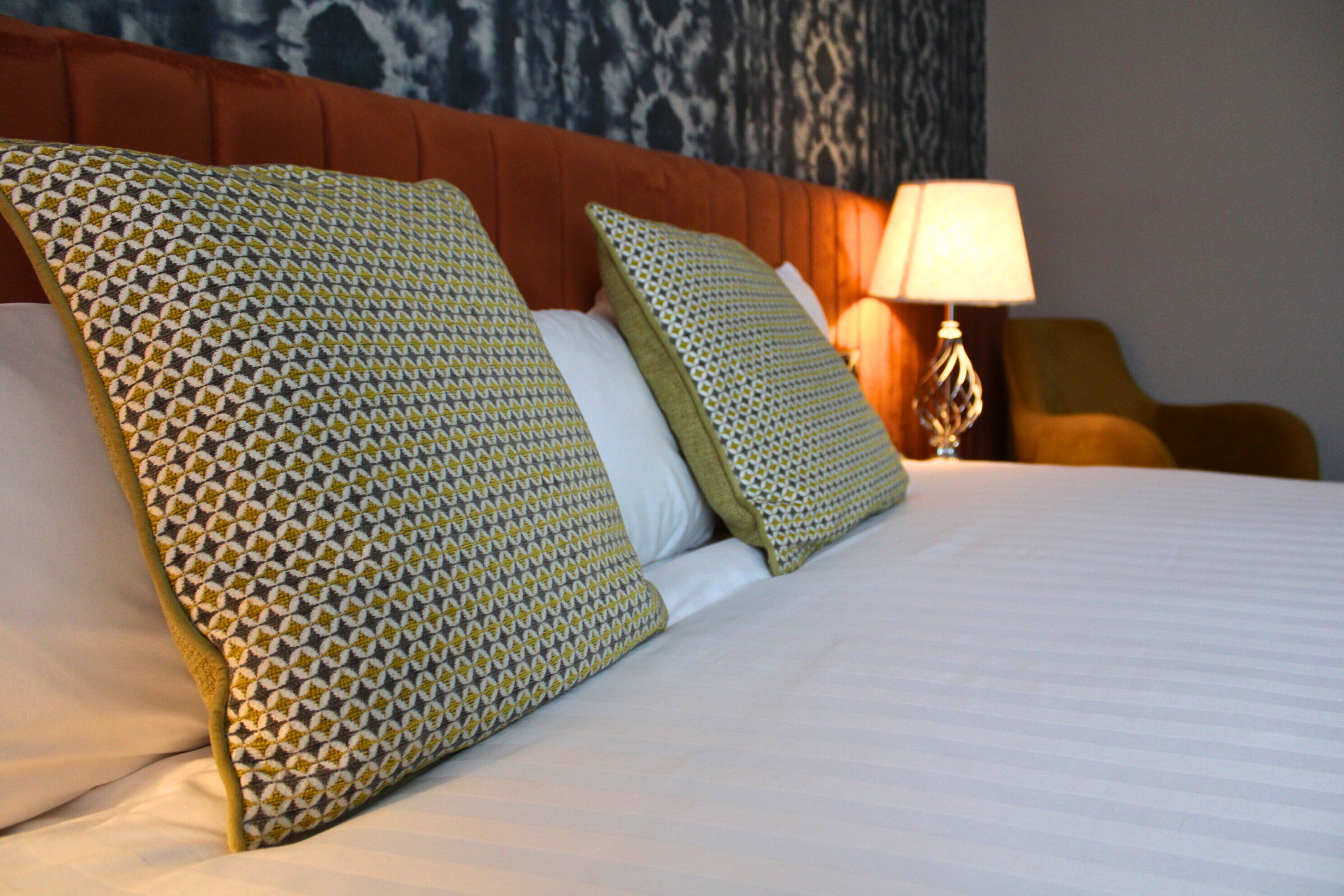 A neatly made bed with patterned pillows and a headboard, a bedside lamp, and a chair in a hotel room.