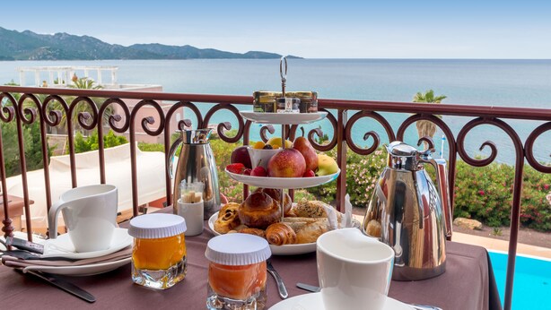 A breakfast setup with a tiered fruit and pastry display, coffee pots, and a scenic ocean view. - Demeure Loredana  (Saint-Florent)