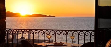 A balcony with a table set for two, overlooking a sunset over the sea.