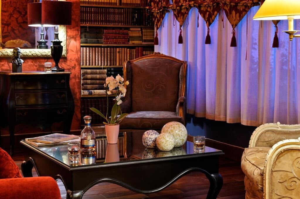 Une pièce chaleureuse avec un fauteuil en cuir, une table basse en verre agrémentée d’objets décoratifs, une bibliothèque remplie de livres et un lustre classique.