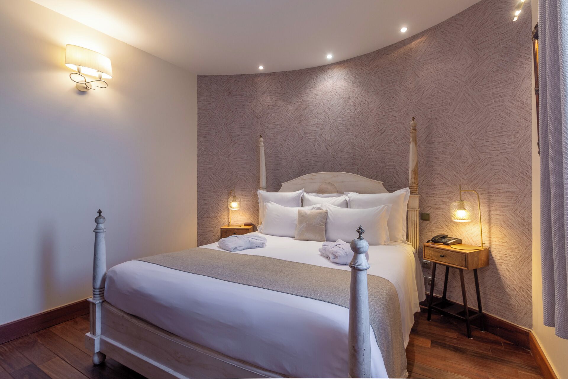 A neatly arranged bedroom with a four-poster bed, two bedside tables with lamps, and a patterned wallpaper.