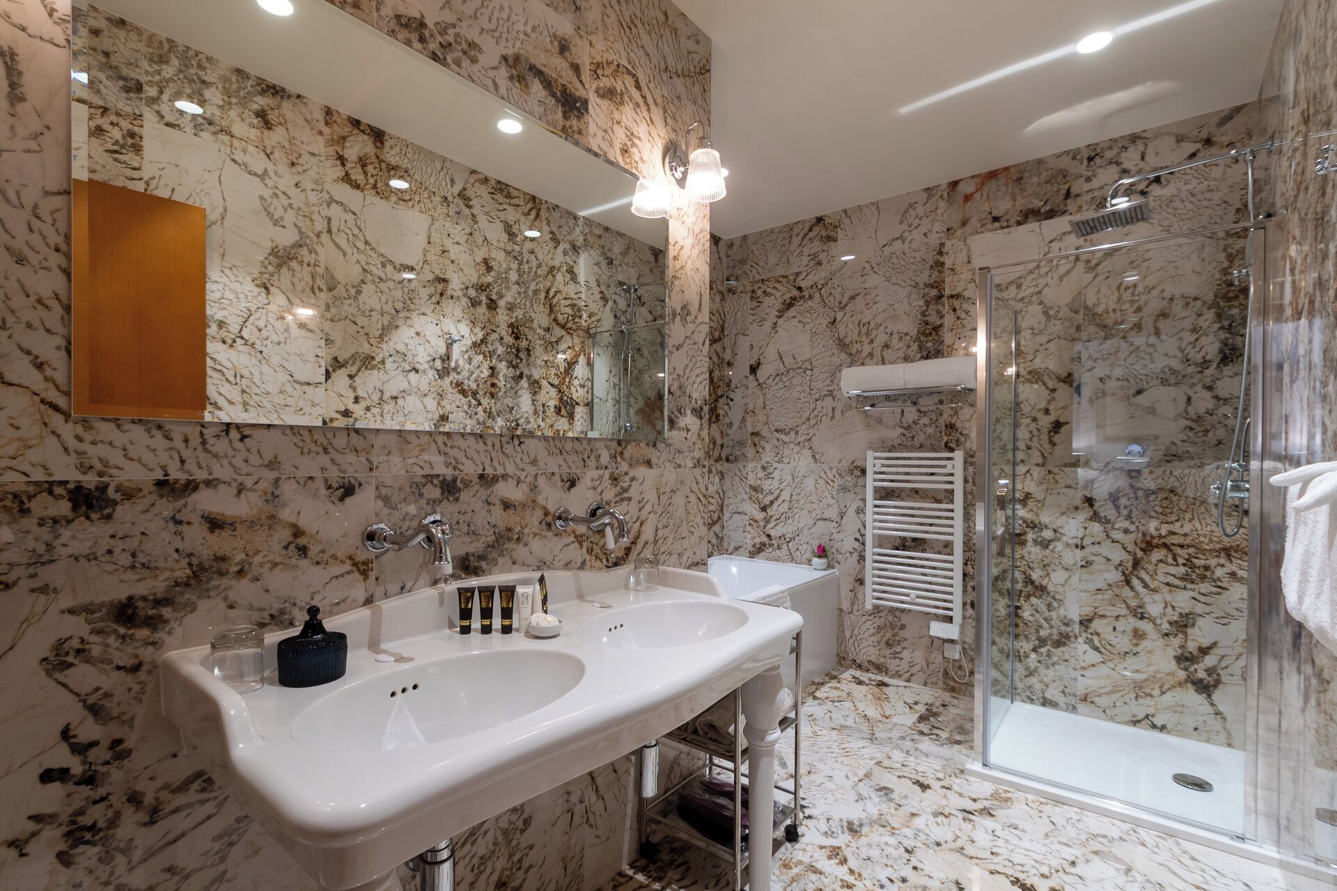 A bathroom with a marble backsplash, a double sink vanity, a glass shower enclosure, and a towel rack.