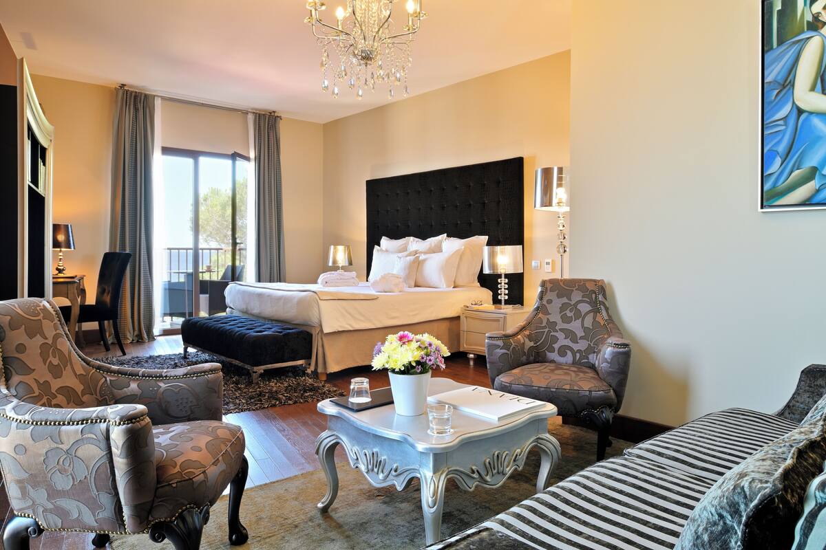 Une chambre d’hôtel avec un grand lit, deux fauteuils, un canapé, une table basse ornée d’un vase de fleurs, un lustre et un tableau accroché au mur.