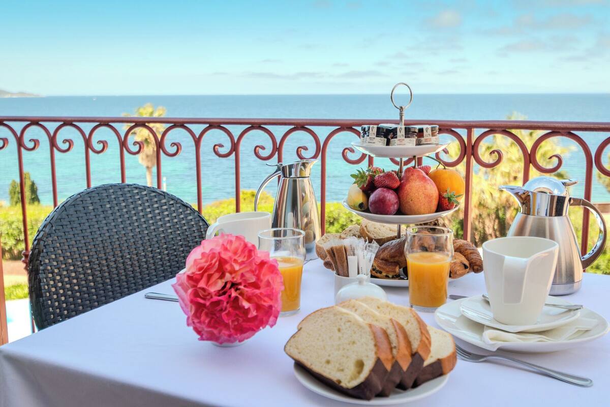 Une table dressée avec des aliments pour le petit-déjeuner, notamment du pain, des fruits et du café, offrant une vue panoramique sur l’océan.
