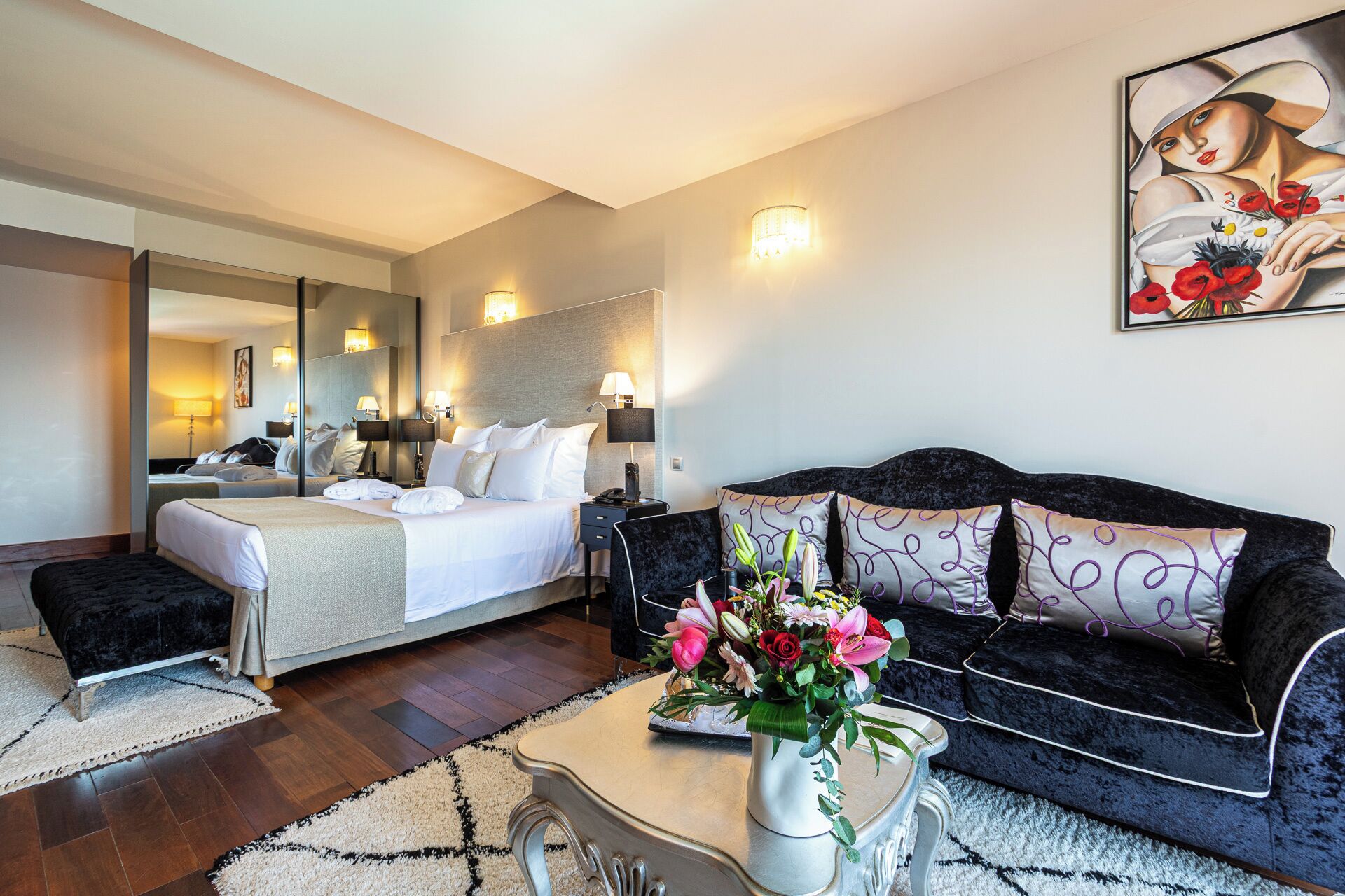 Une chambre d’hôtel avec un lit, un canapé, une table basse agrémentée de fleurs, un miroir et des lampes fixées au mur.