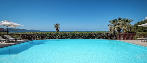 A clear blue pool with lounge chairs and umbrellas, surrounded by greenery and a view of the sea.