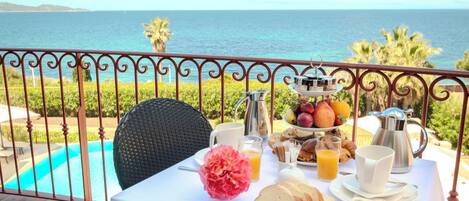 A breakfast setup with a table, drinks, and a fruit tray on a balcony with a view of the sea.