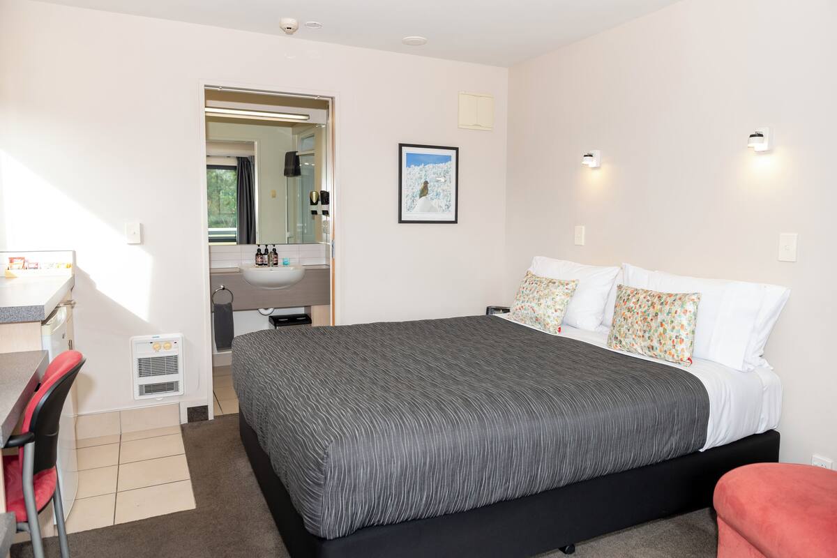 A bedroom with a large bed, a desk with a chair, a mirror, a sink, and a framed picture on the wall.