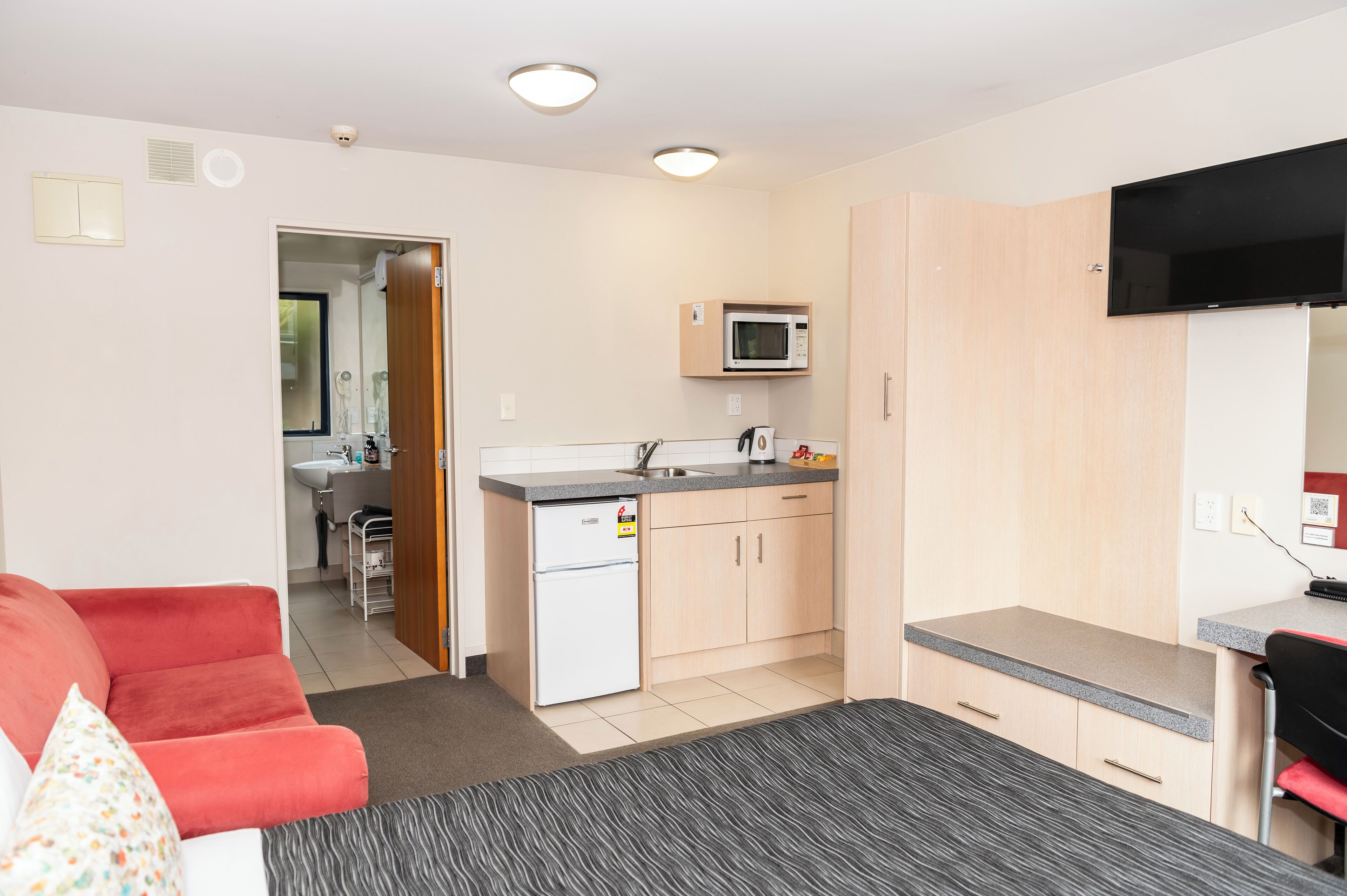 A compact living space with a kitchenette, a red sofa, a small dining table, and a TV mounted on the wall.
