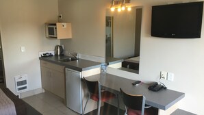 Cocina compacta con microondas, lavamanos y un refrigerador pequeño. Hay una zona de comedor con una silla y una mesa, y un televisor de pantalla plana montado en la pared.