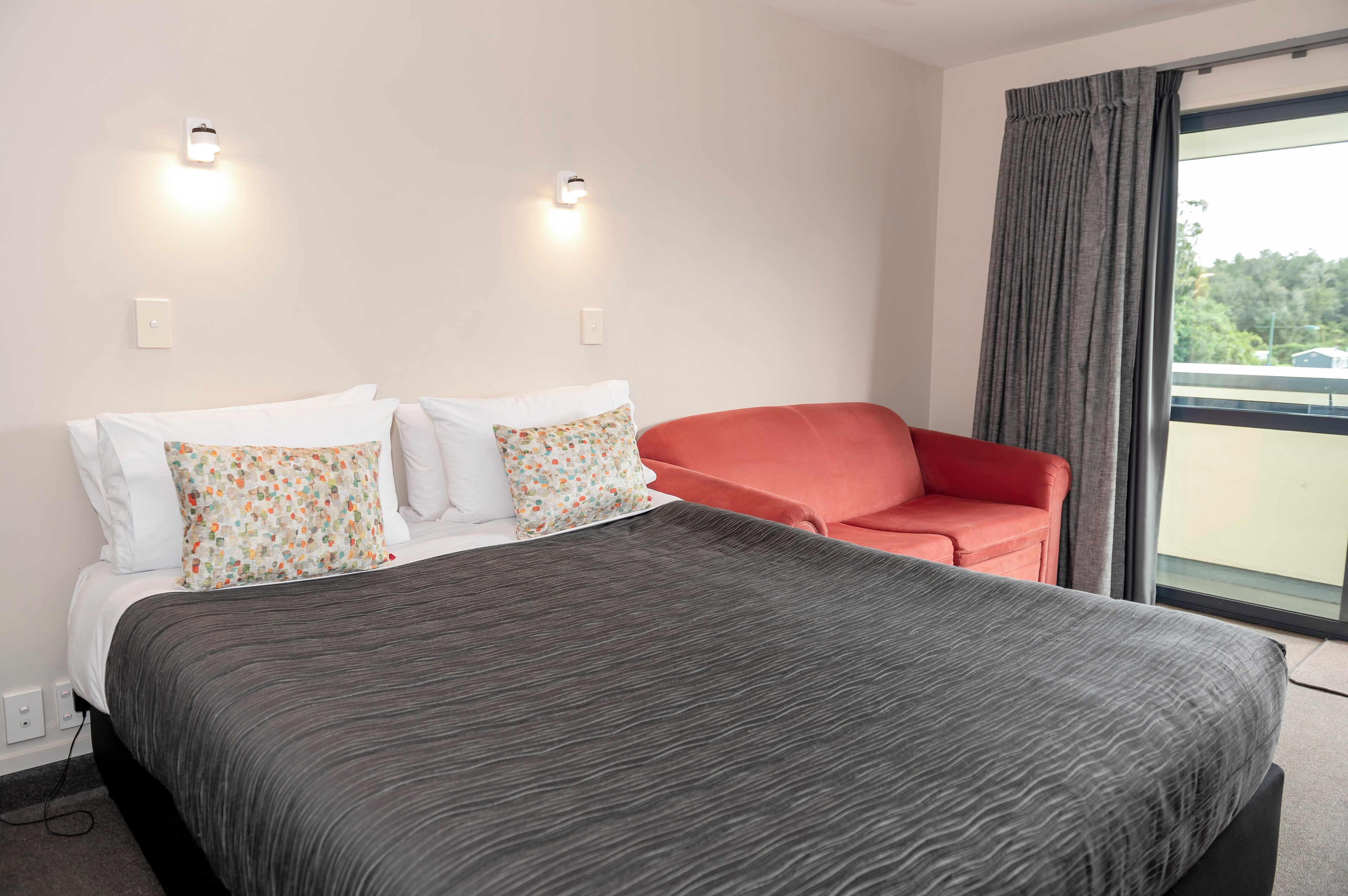 Une chambre d’hôtel avec un grand lit, deux oreillers, une chaise rouge et une fenêtre avec des rideaux.