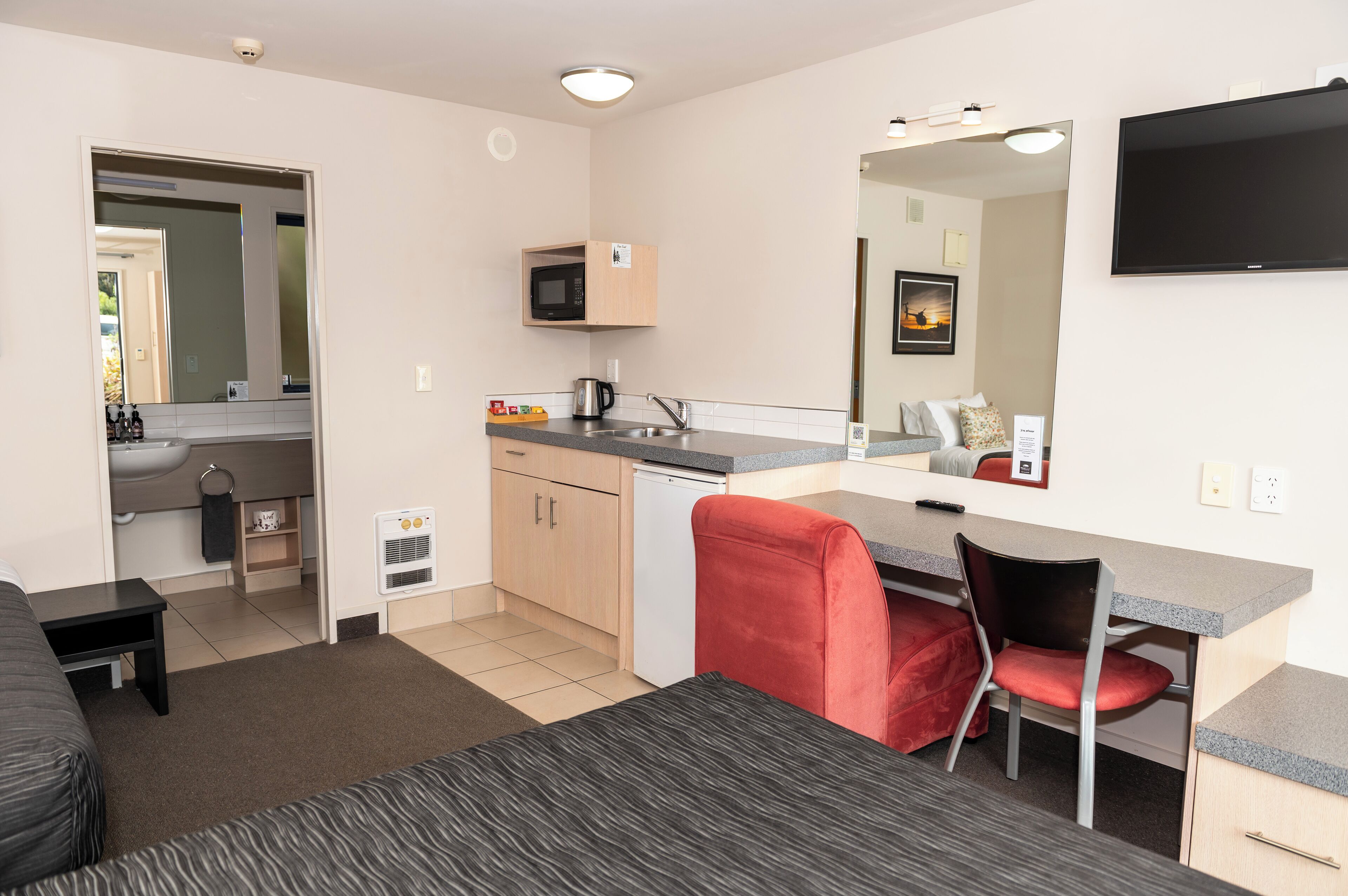 Habitación de hotel compacta con kitchenette, zona de comedor con una silla roja, un pequeño sofá y un televisor de pantalla plana montado en la pared.