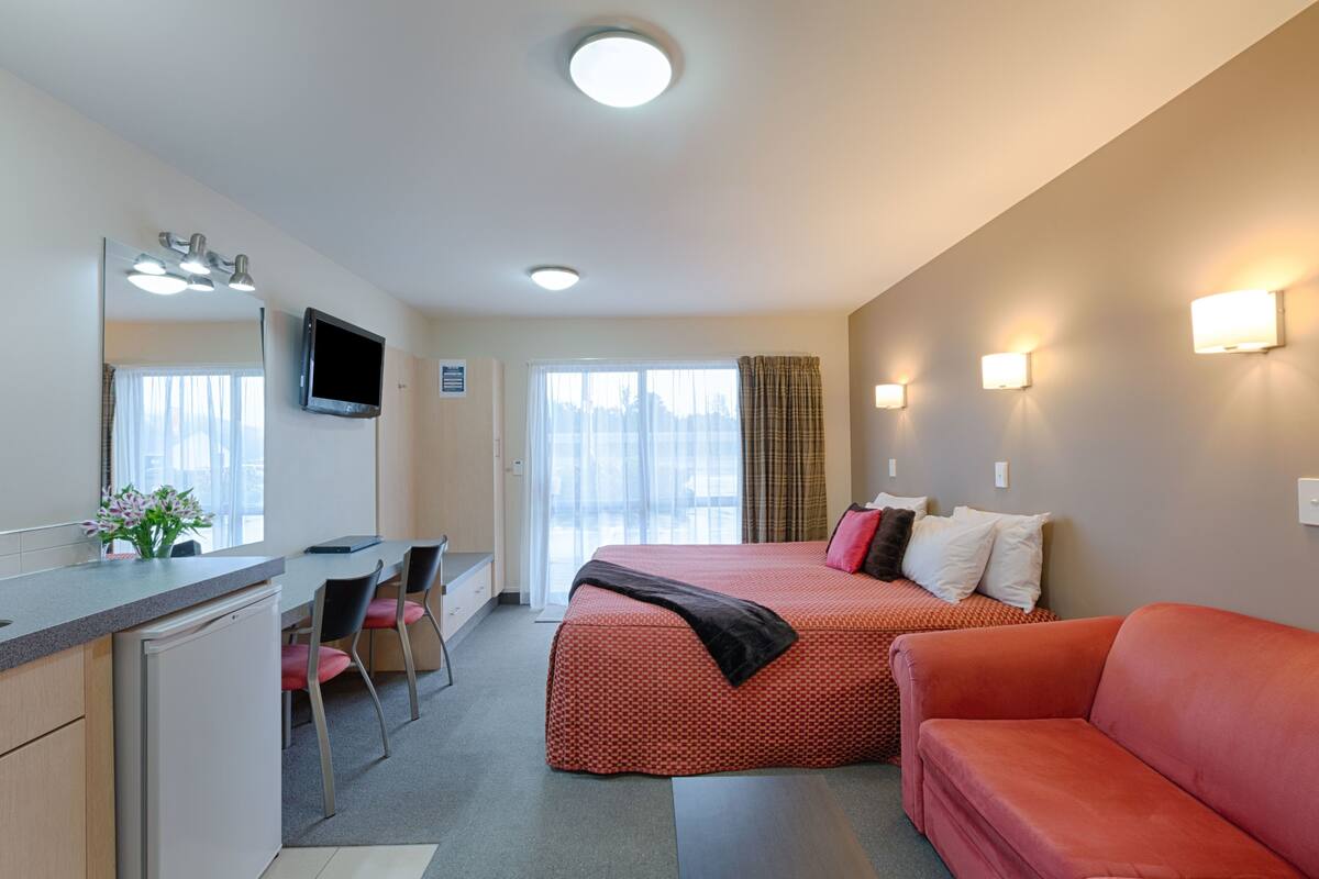 A hotel room with a bed, a desk, a chair, a sofa, a TV, and a window with curtains.