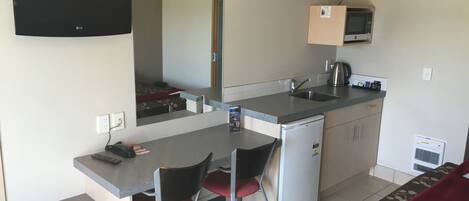 Una kitchenette compacta con microondas, lavamanos y refrigerador pequeño. Hay una zona de comedor con dos sillas y una mesa.