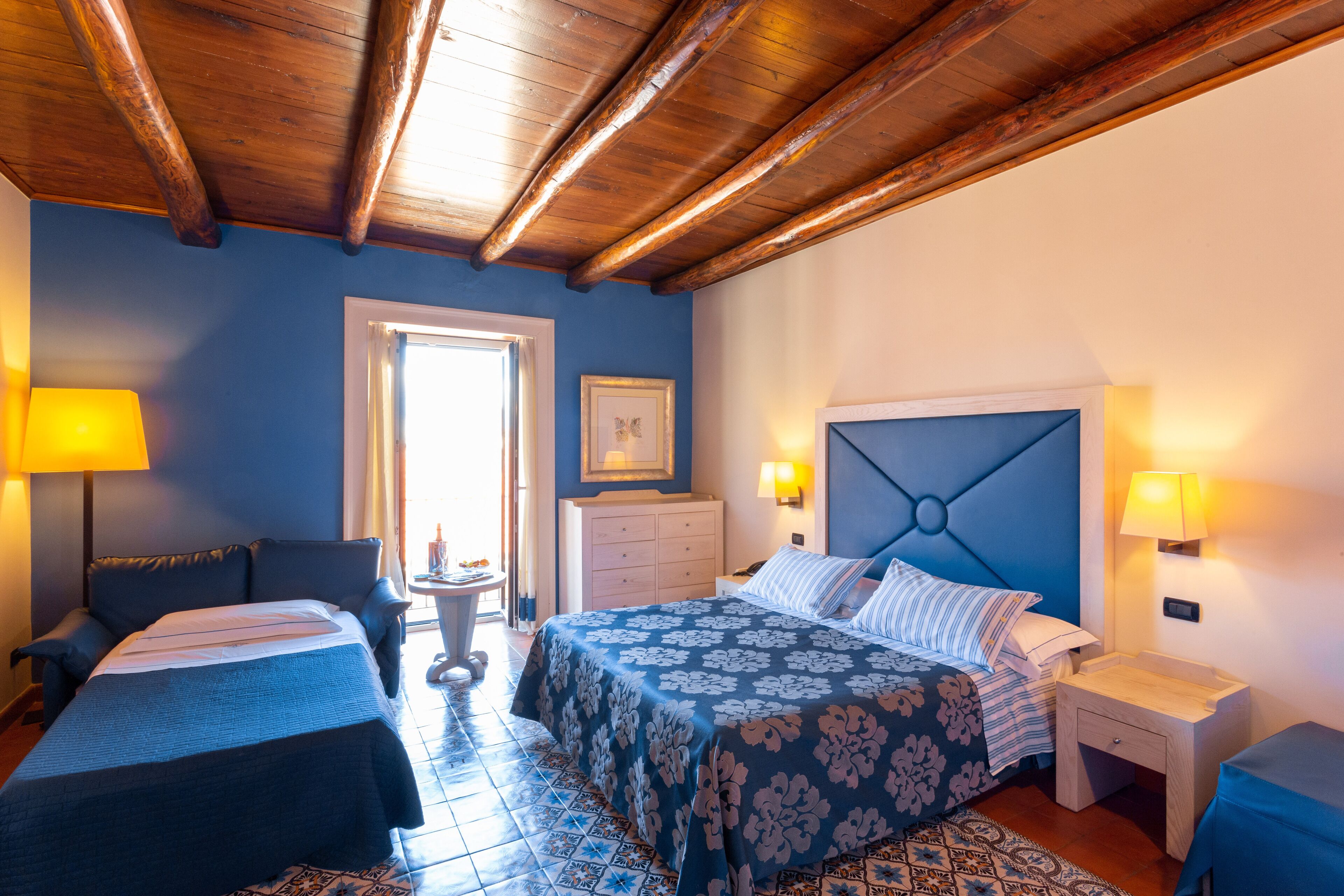 A hotel room with two beds, a wooden ceiling, a blue wall, and a tiled floor.