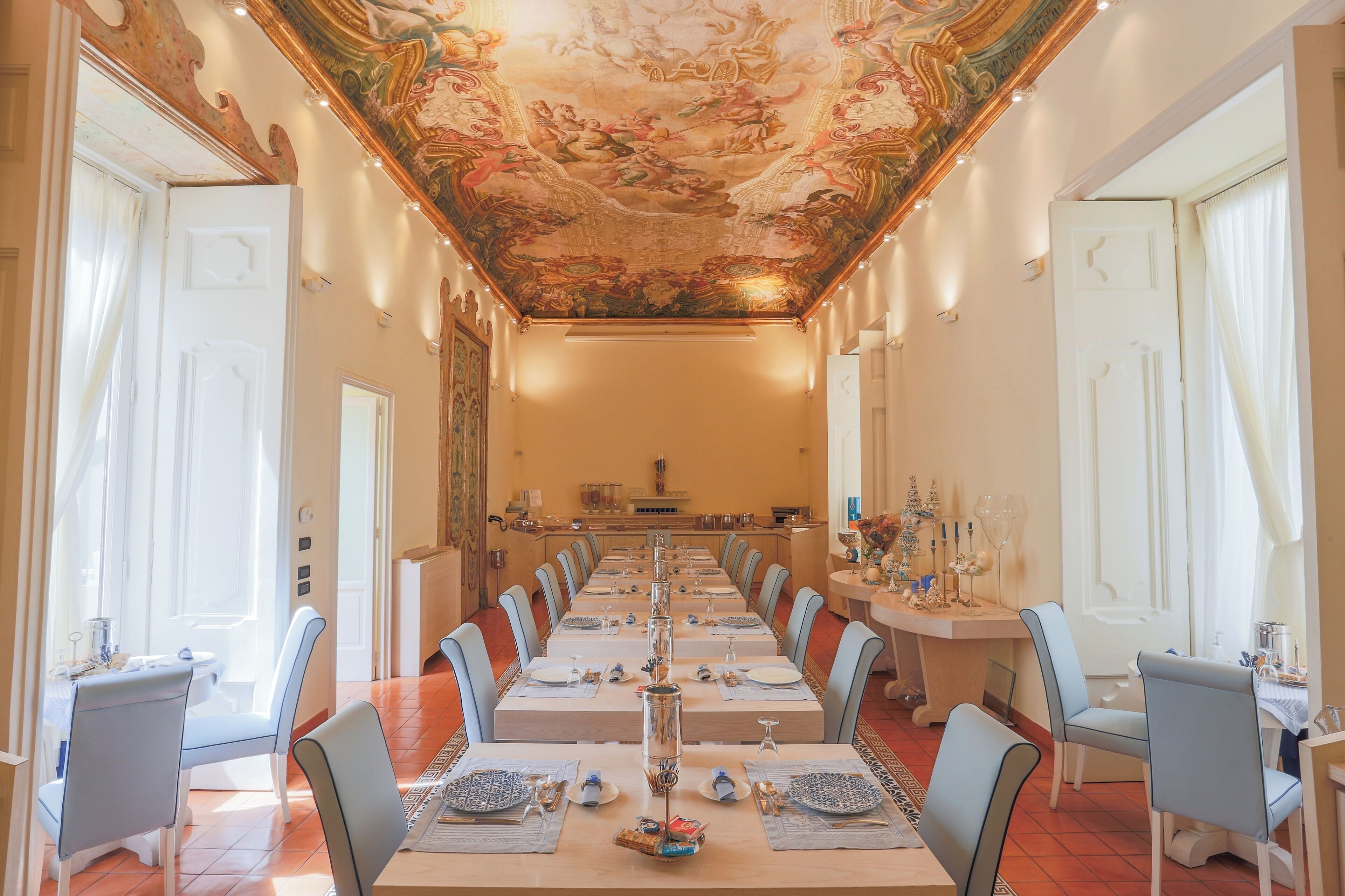 A long dining table set for a formal meal, with chairs on both sides, in a room with ornate ceiling artwork and a chandelier.