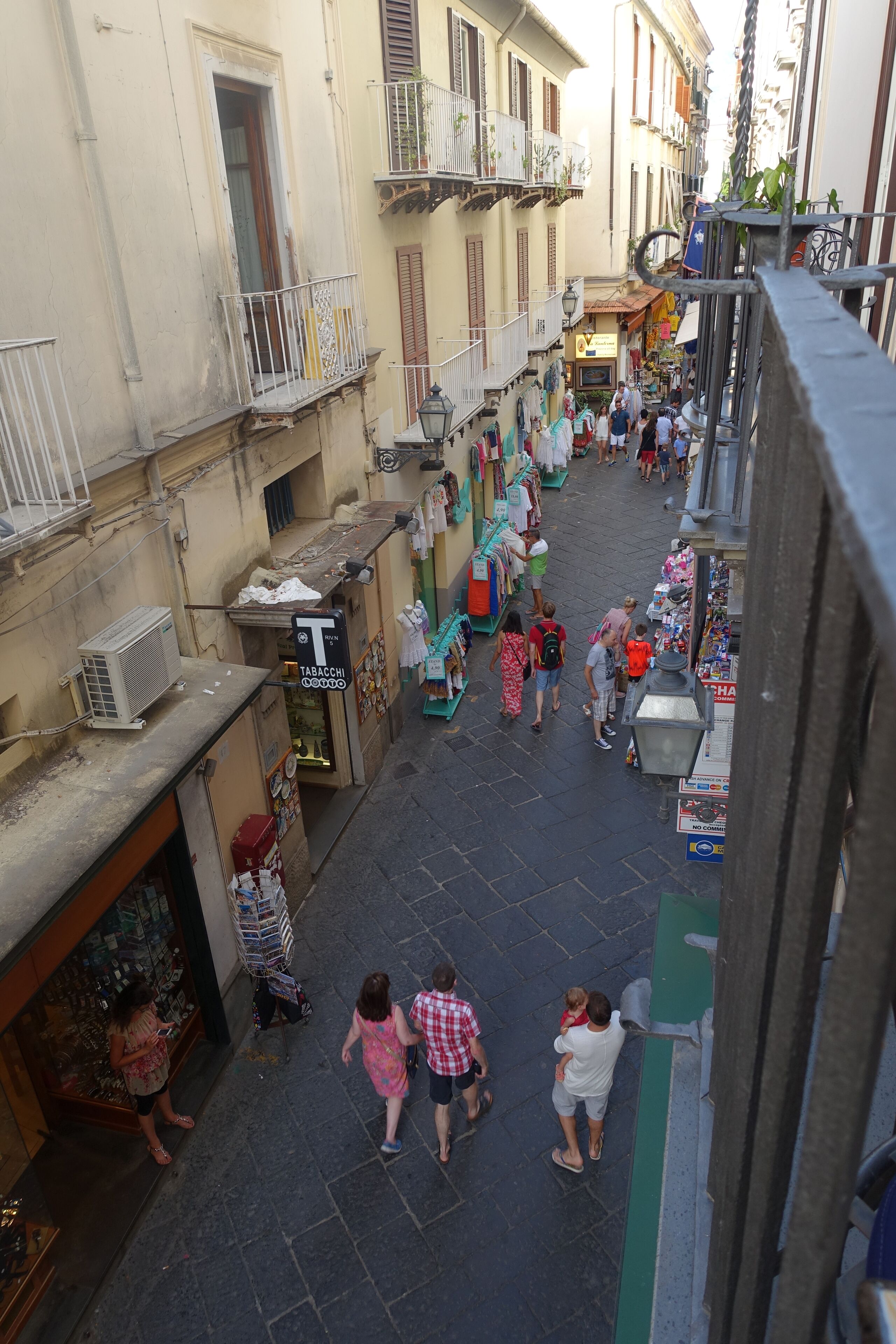 A narrow street with shops on either side, balconies, and a few pedestrians.