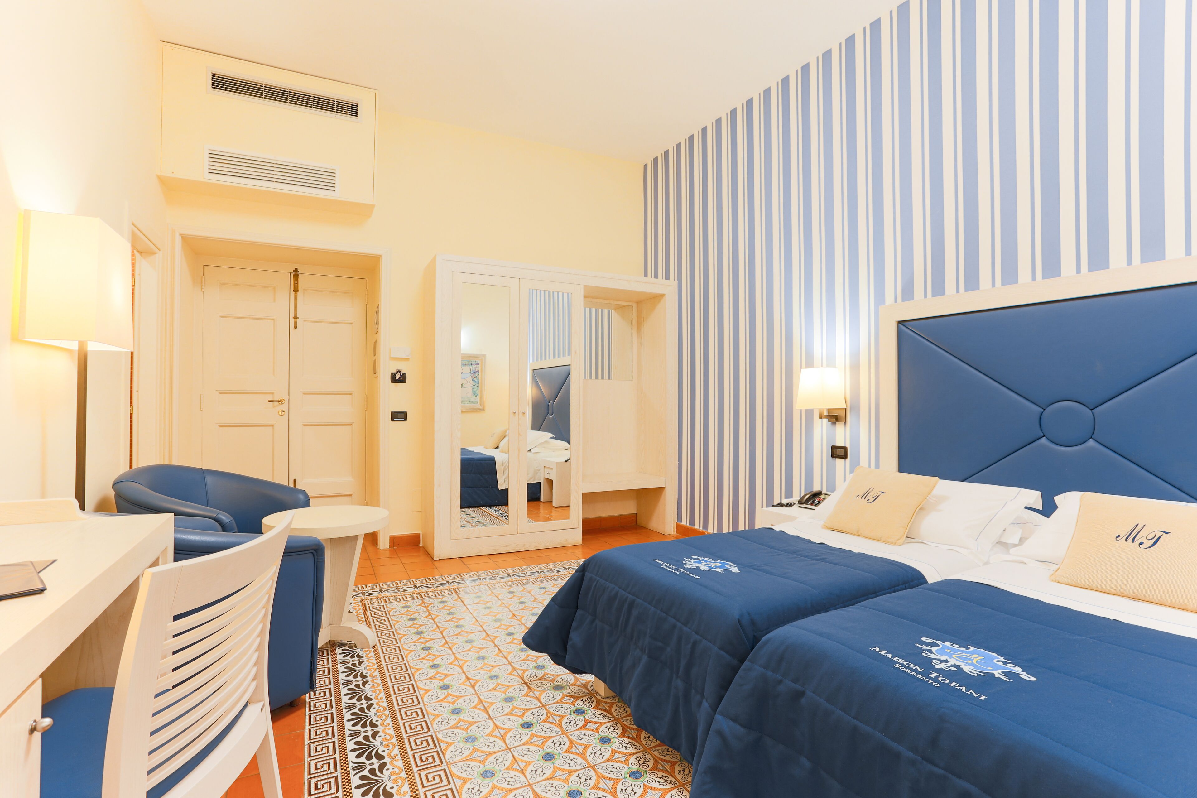 A hotel room with two beds, a desk, a chair, a mirror, and a striped wall.