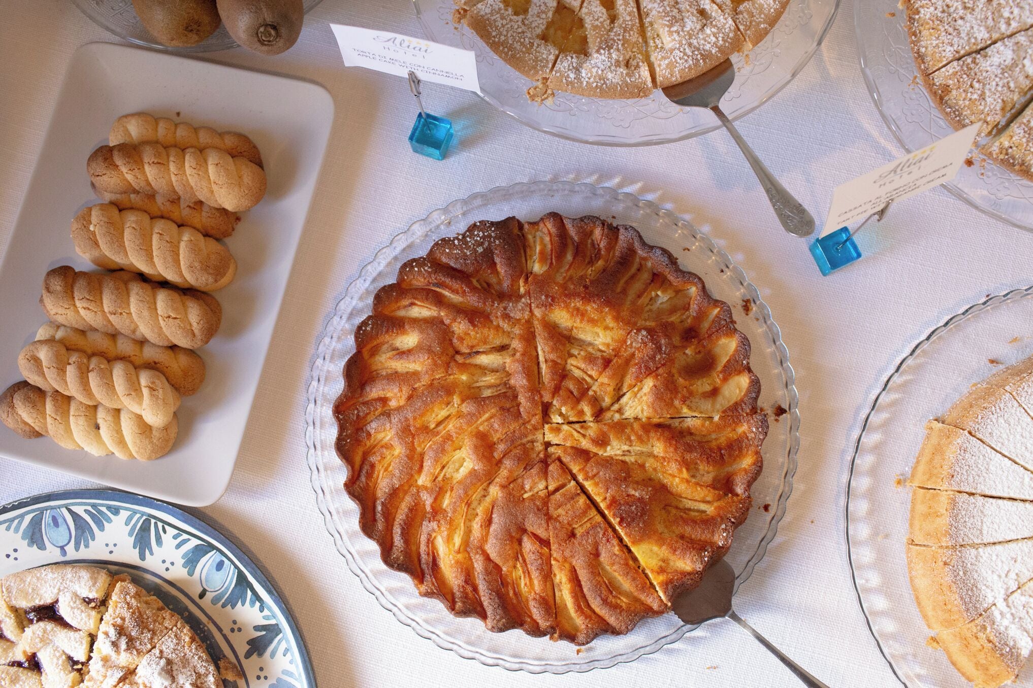 A table with various baked goods, including a pie with a lattice crust, a plate of braided bread, and a round cake with a dusting of powdered sugar.