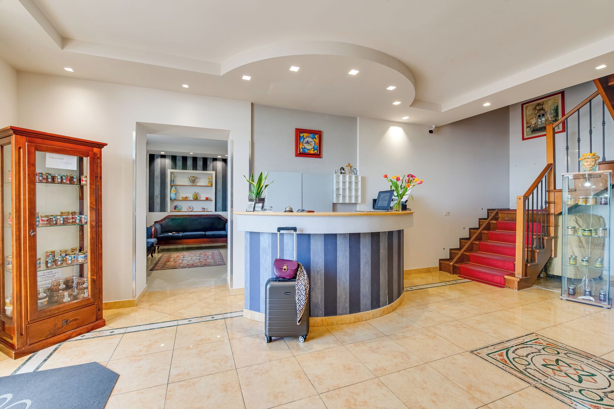 A hotel lobby with a reception desk, a staircase, a wooden cabinet with jars, and a tiled floor.