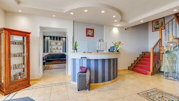 A hotel lobby with a reception desk, a staircase, a wooden cabinet with jars, and a tiled floor.