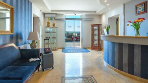 A hotel lobby with a sofa, a reception desk, a mirror, a lamp, a suitcase, and a view through the glass doors. - Hotel Aliai (Sciacca)