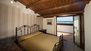 A bedroom with a wooden ceiling, a bed with striped bedding, a nightstand, a framed picture on the wall, and a balcony with a view of the sea. - Hotel Aliai (Sciacca)