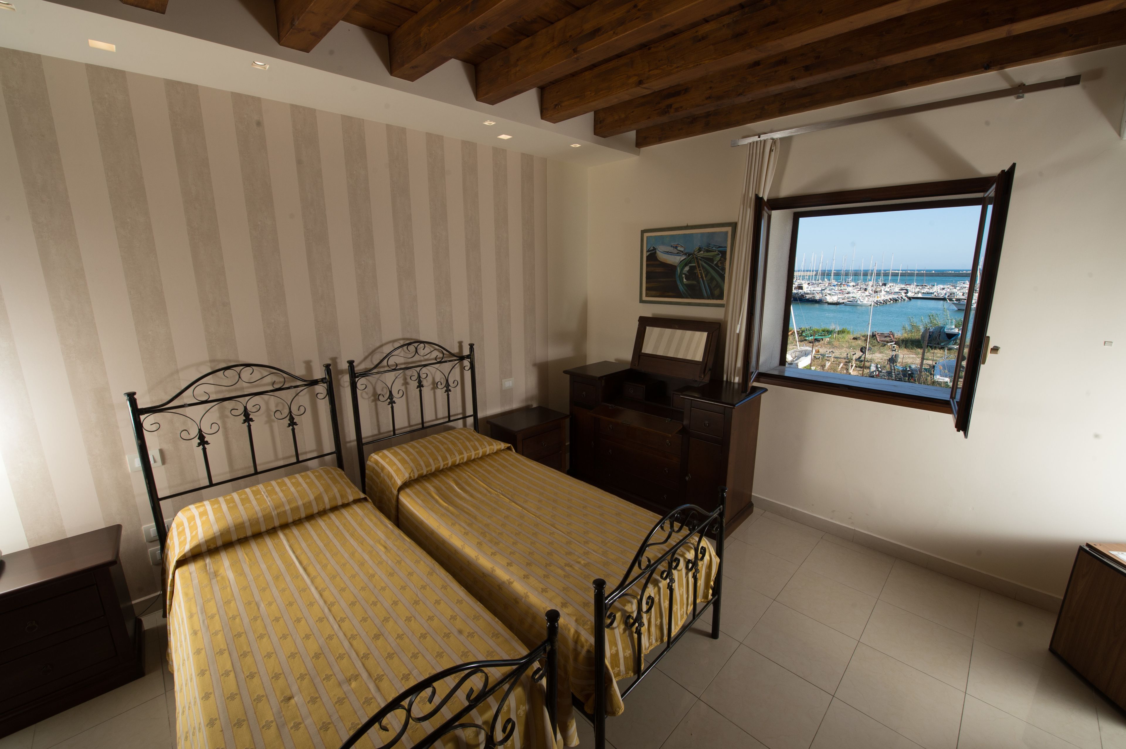 A bedroom with two beds, a wooden ceiling, a window with a view of a marina, and striped wallpaper.