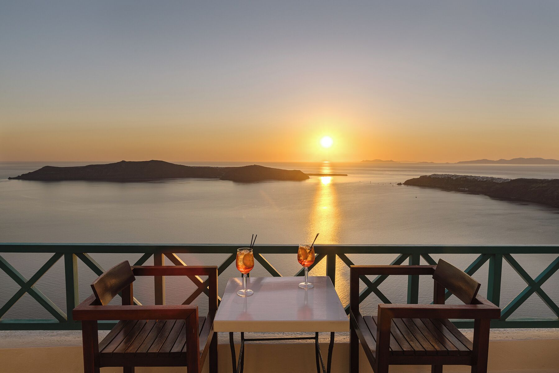 A table with two chairs and two drinks, overlooking a sunset over a calm sea with distant landforms.