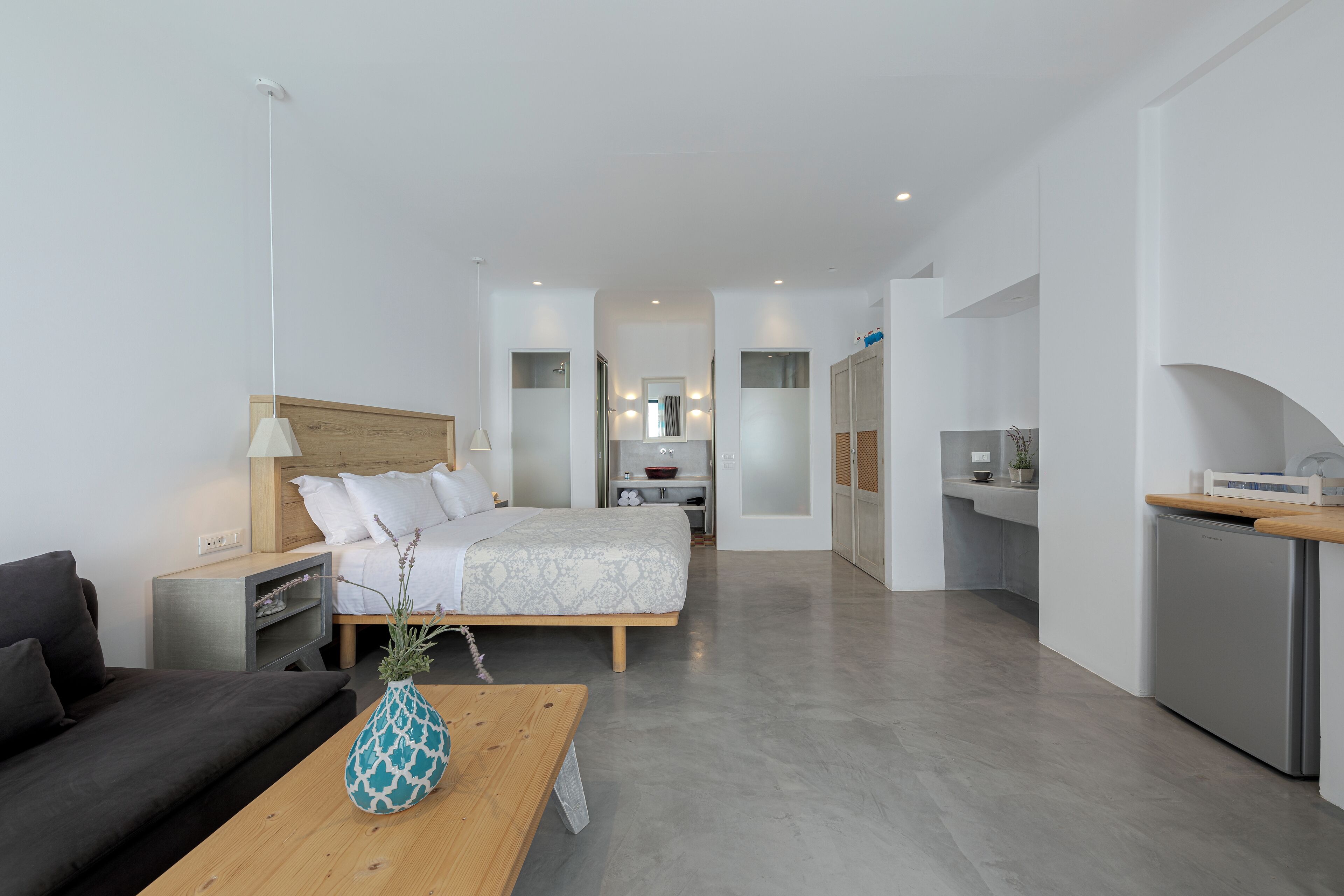 A modern hotel room with a bed, a sofa, a dining table with a vase, a kitchen area, and a bathroom visible through an archway.
