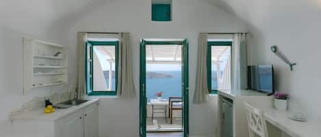 A compact room with a bed, a small kitchenette, a dining area with a table and chairs, and a view of the sea through open windows.