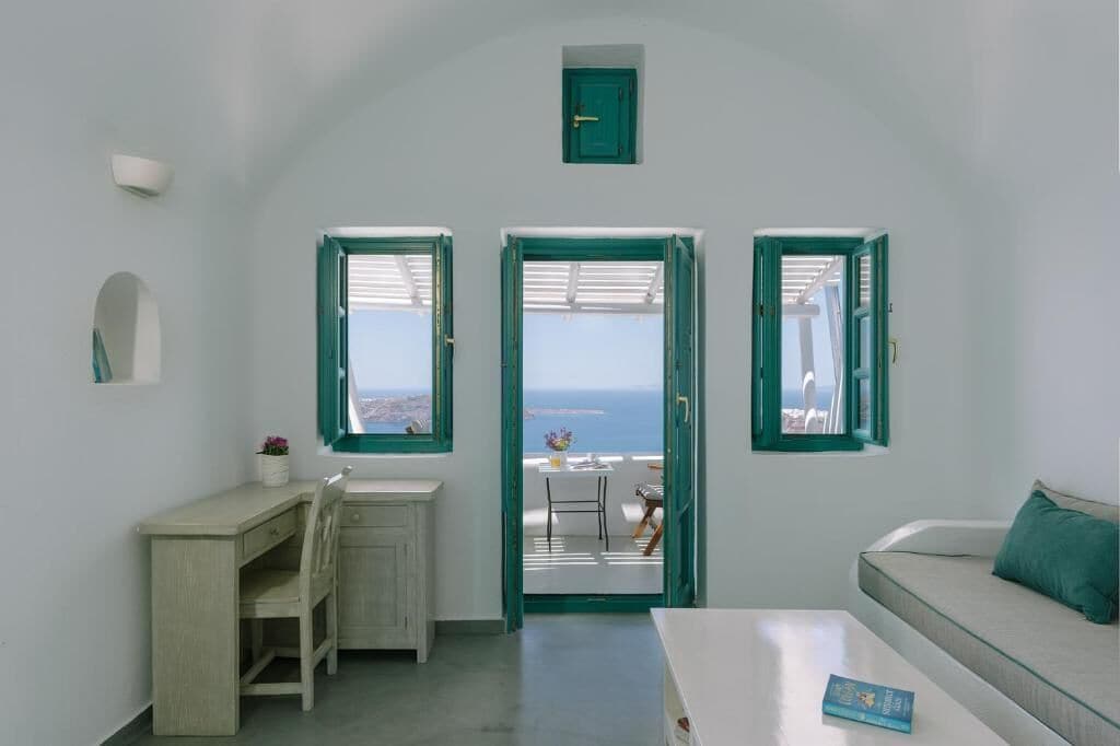 Une pièce comprenant un bureau, une chaise, un canapé et offrant une vue sur la mer grâce à de grandes fenêtres ouvertes.