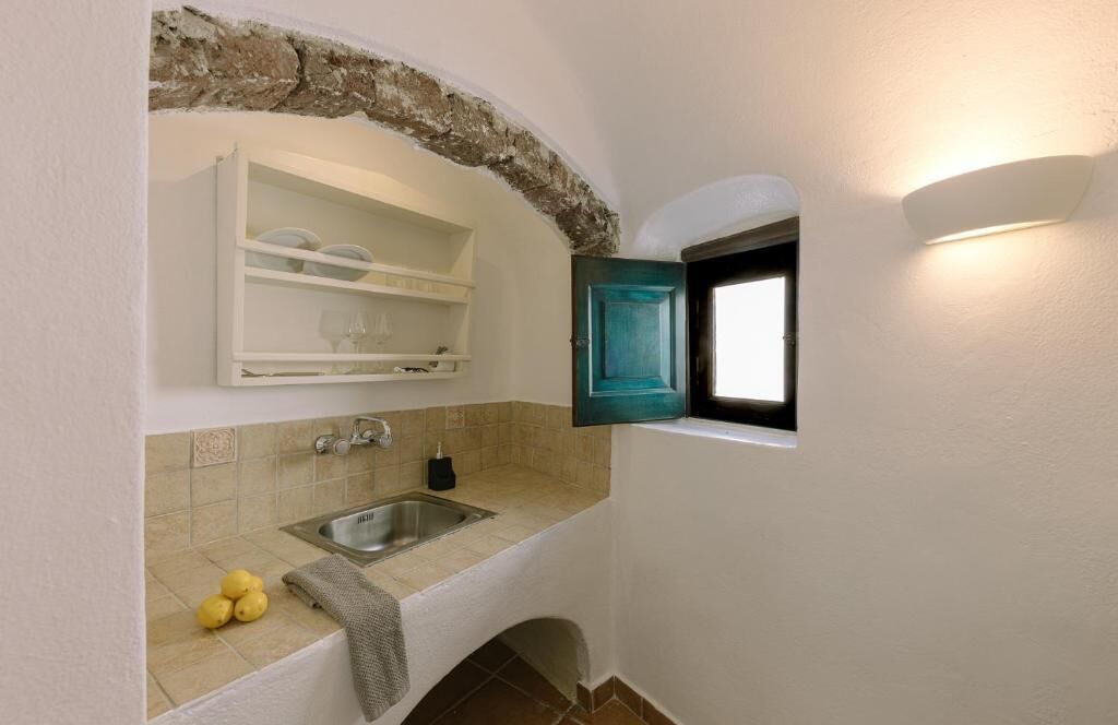 A narrow kitchen with a built-in sink, a window with a teal door, and a shelf above the sink.