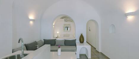 Un interior moderno y minimalista con una zona de cocina, un espacio de estar con sofá y cojines, y una pequeña mesita con un florero.