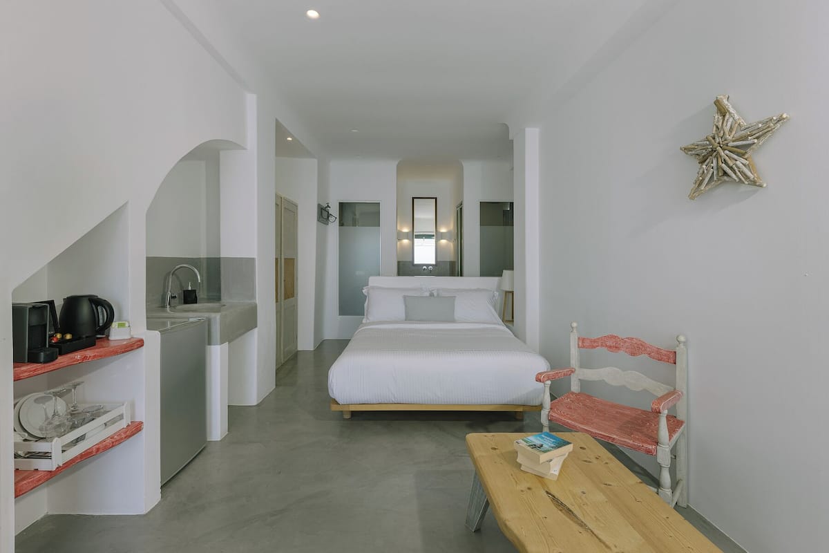 Habitación de hotel con cama, un banco de madera, una mesita con libros, una kitchenette y un baño visible a través de un arco.