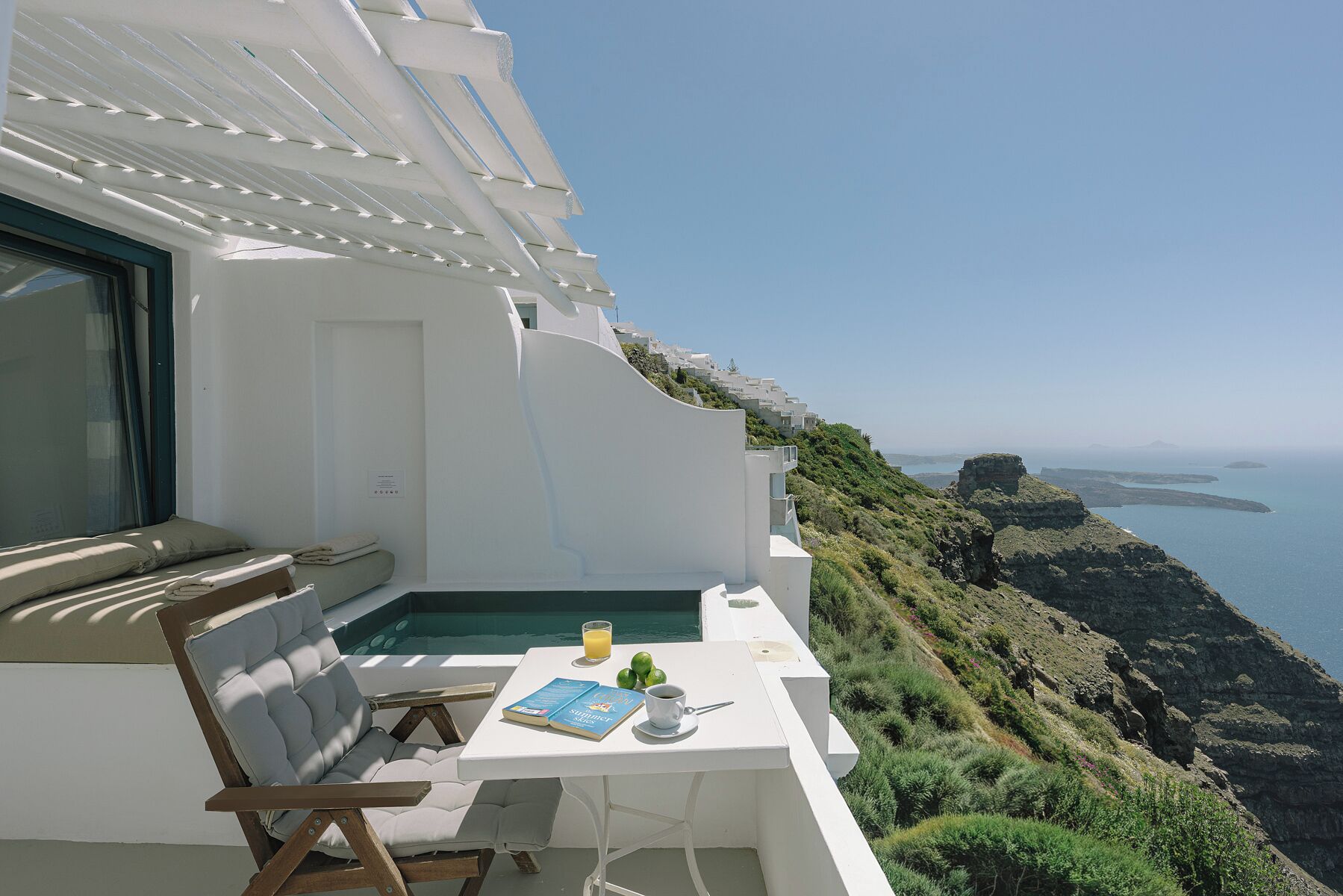 A balcony with a table, a chair, a book, a glass of juice, and a view of the sea and cliffs.