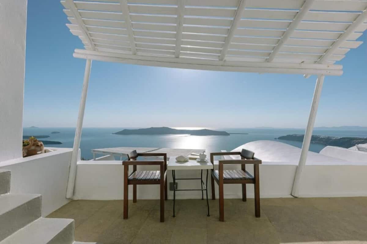 Una terraza con una pérgola blanca, dos sillas de madera, una mesa y vistas al mar y a las islas.