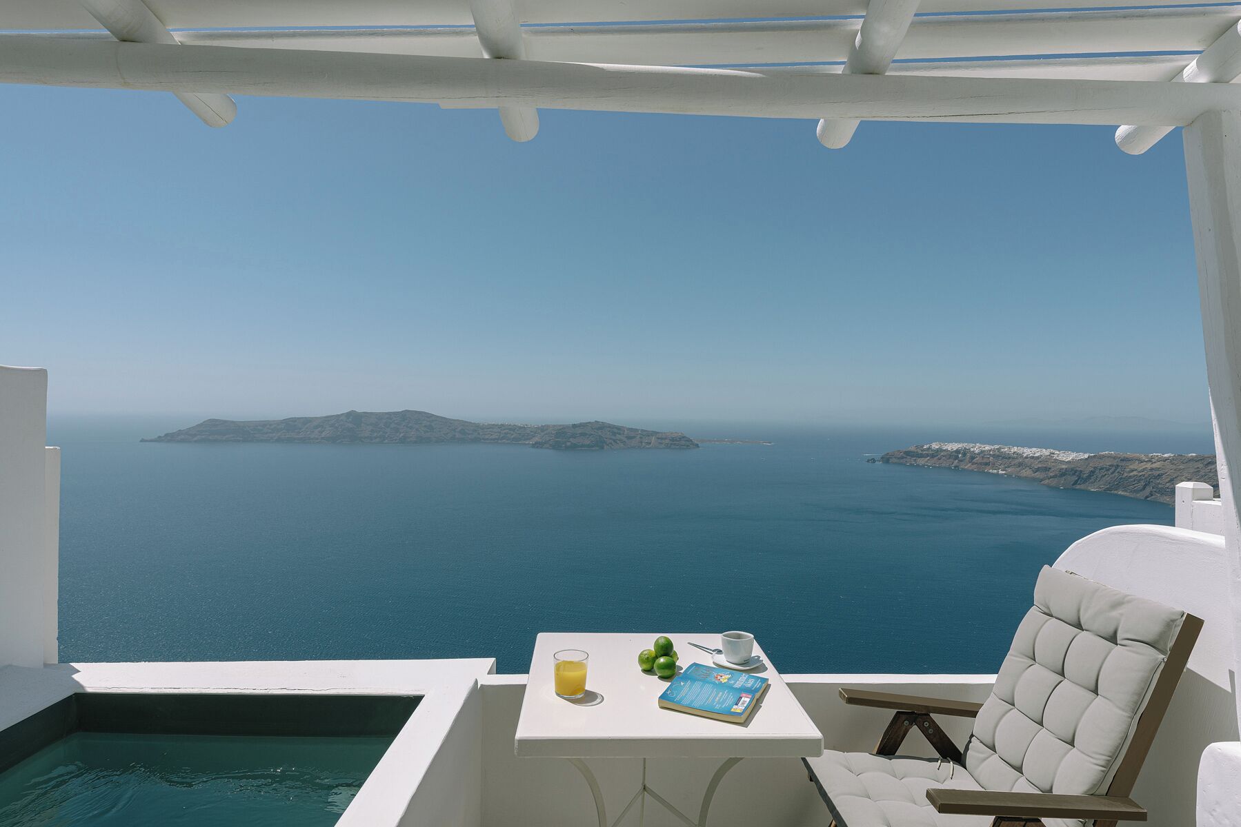 Un balcón con vistas al mar, una mesa con un vaso de zumo de naranja, un libro y una silla.