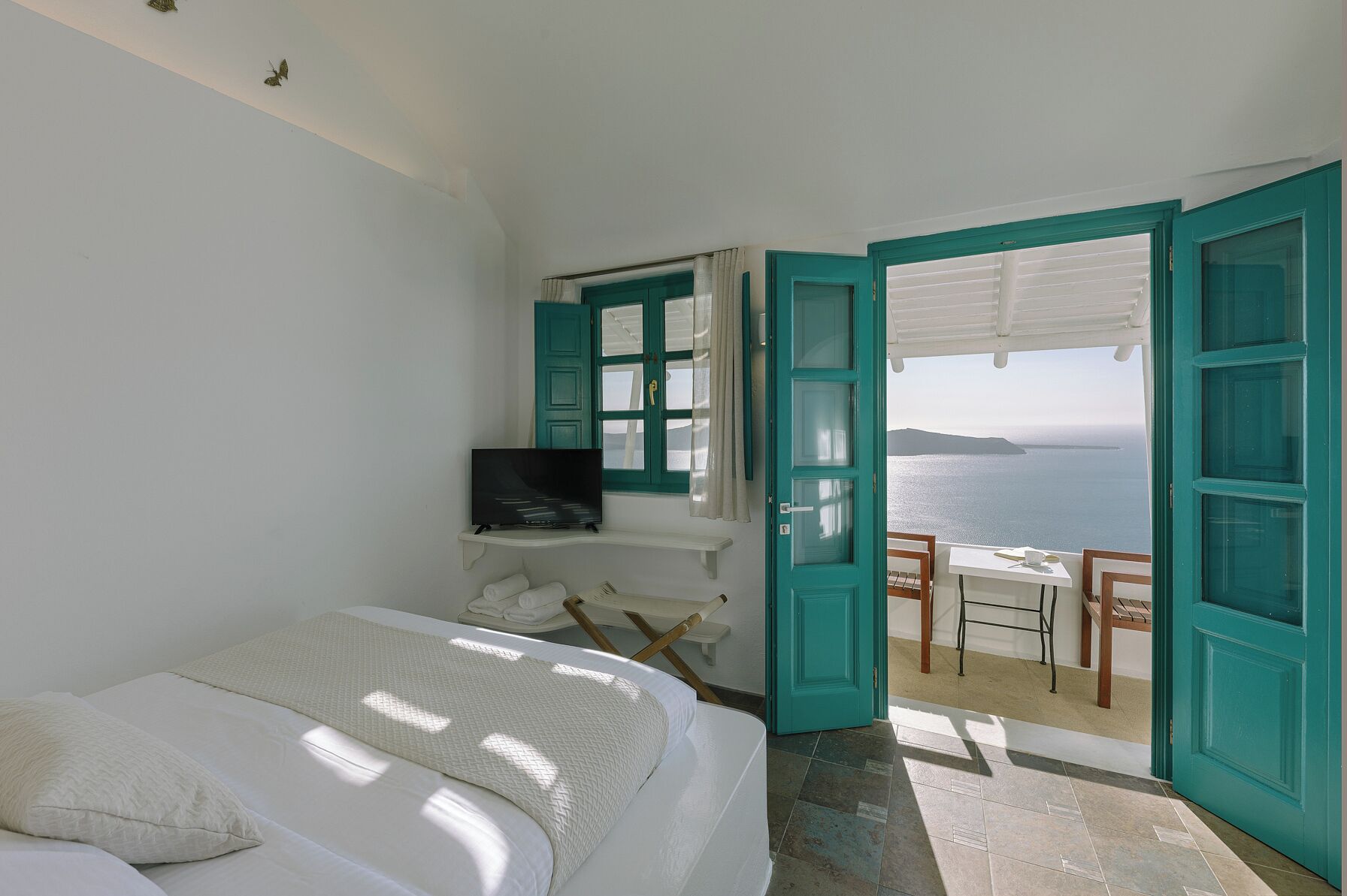 A bedroom with a bed, a TV, a balcony with a table and chairs, and a view of the sea.