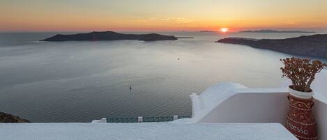 A coastal view with a sunset over calm waters, a white building, and a potted plant on a balcony.