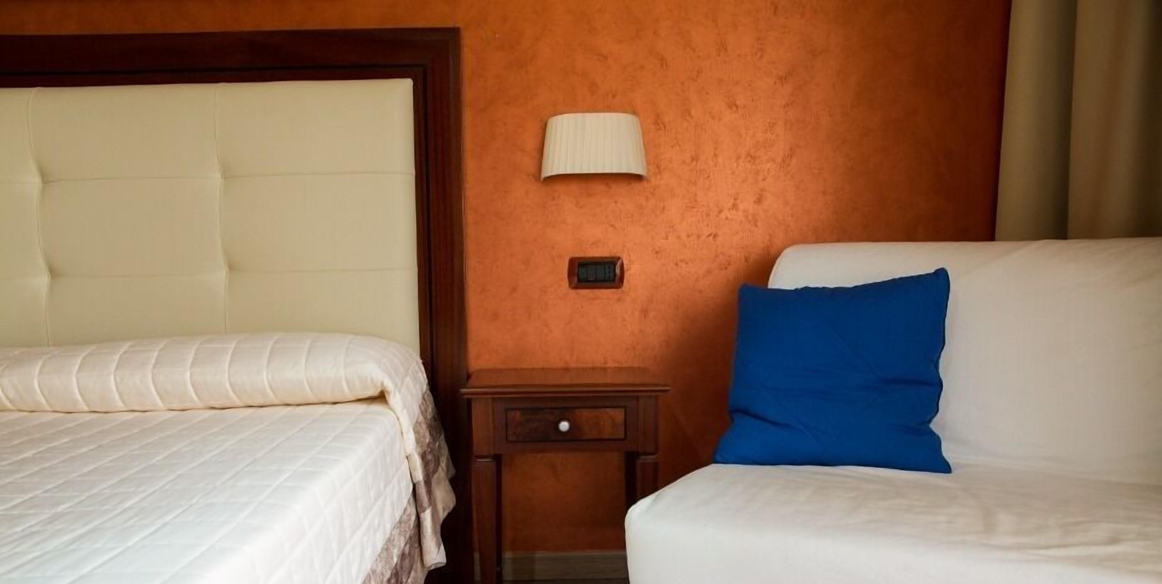 Habitación de hotel con dos camas, una mesita de noche, una lámpara y un termostato en la pared.